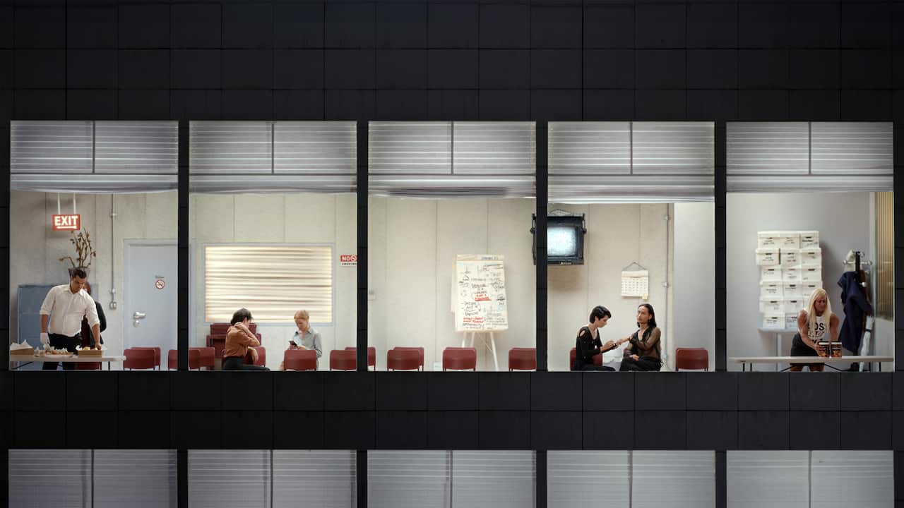 The view through wall of office windows shows a series of people sitting and standing. The lighting suggests is might be after dark.