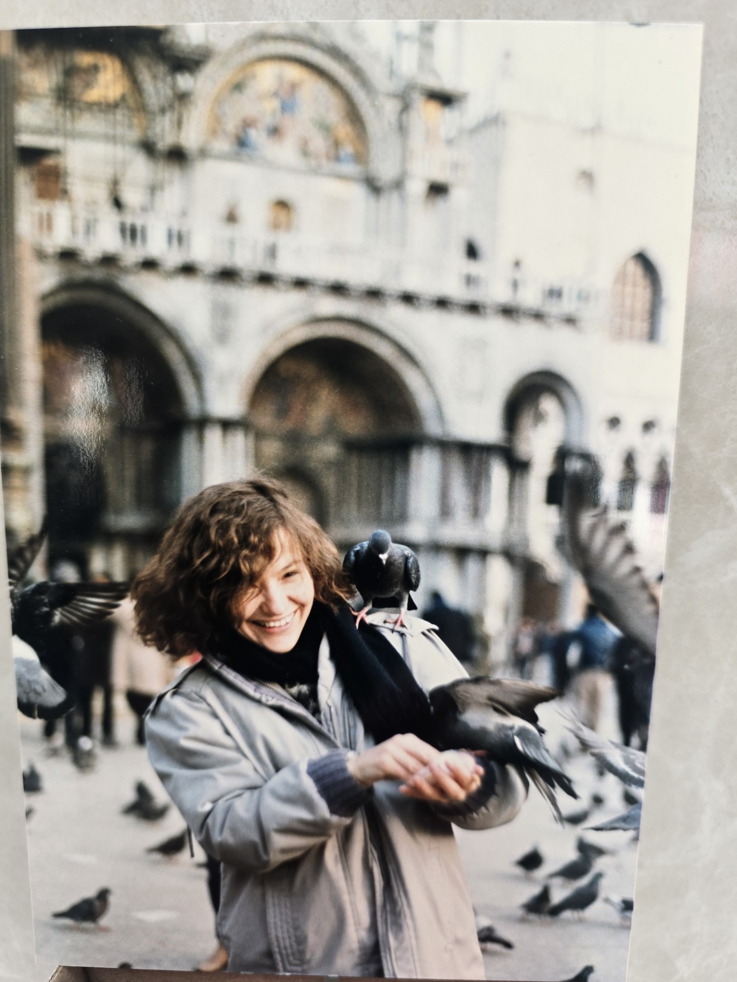 A woman smiles while feeding pigeons in a European square, with birds perched on her shoulders and arms.
