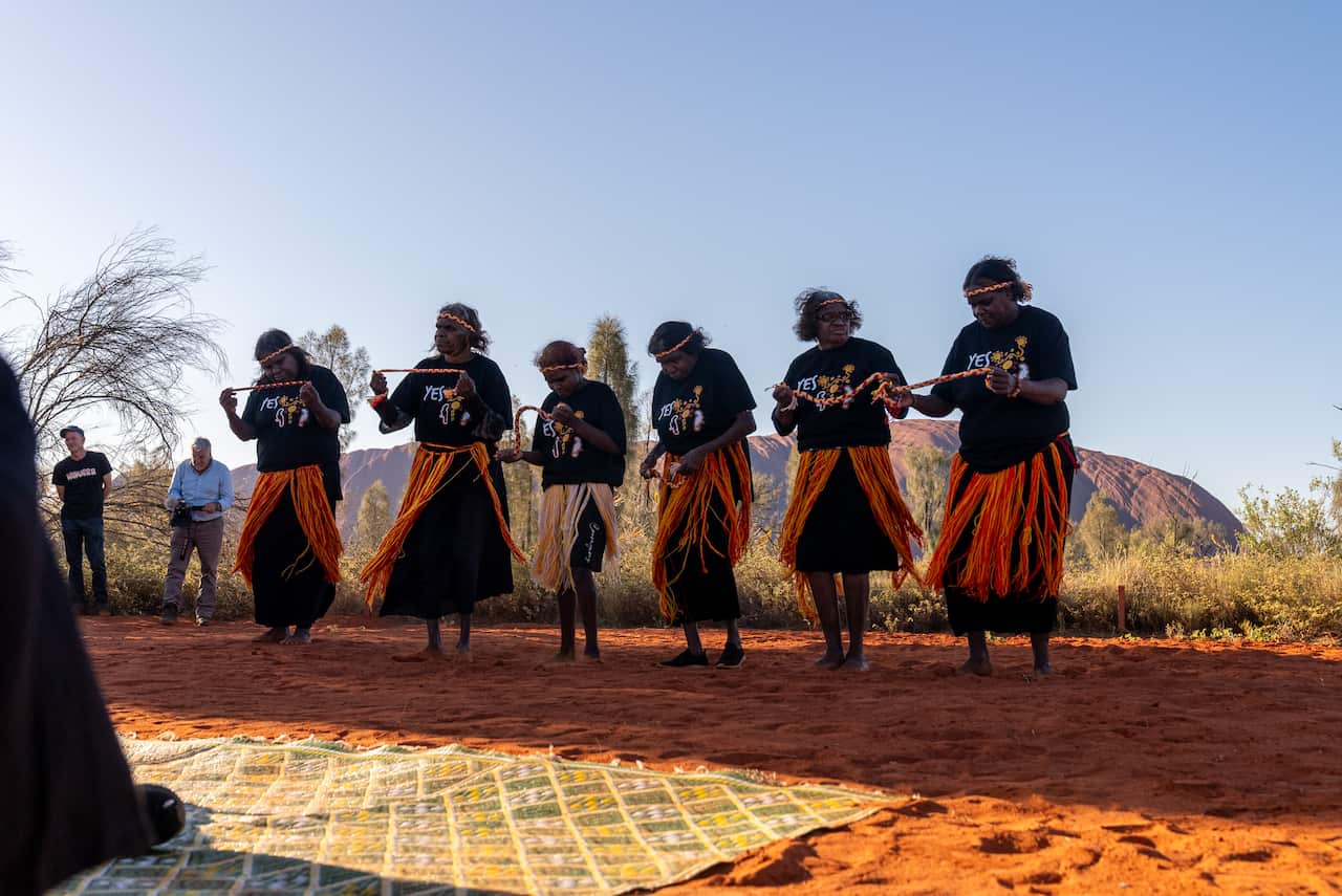 dancers in black red and gold Aboriginal colours perform in the red dirt surrounding uluru