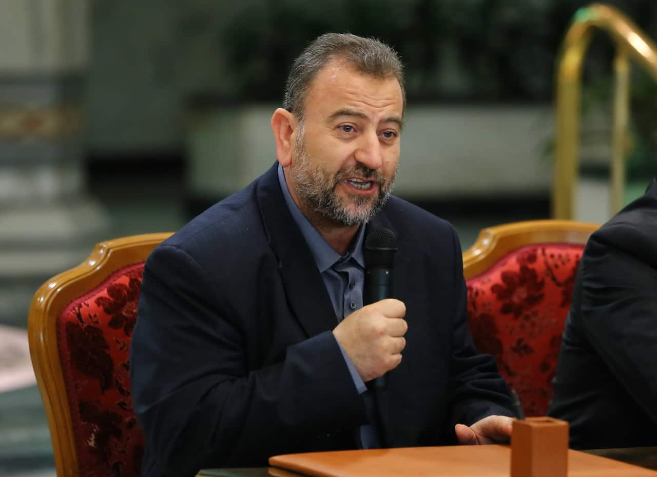 A man with a microphone sitting on a red chair behind a brown desk.