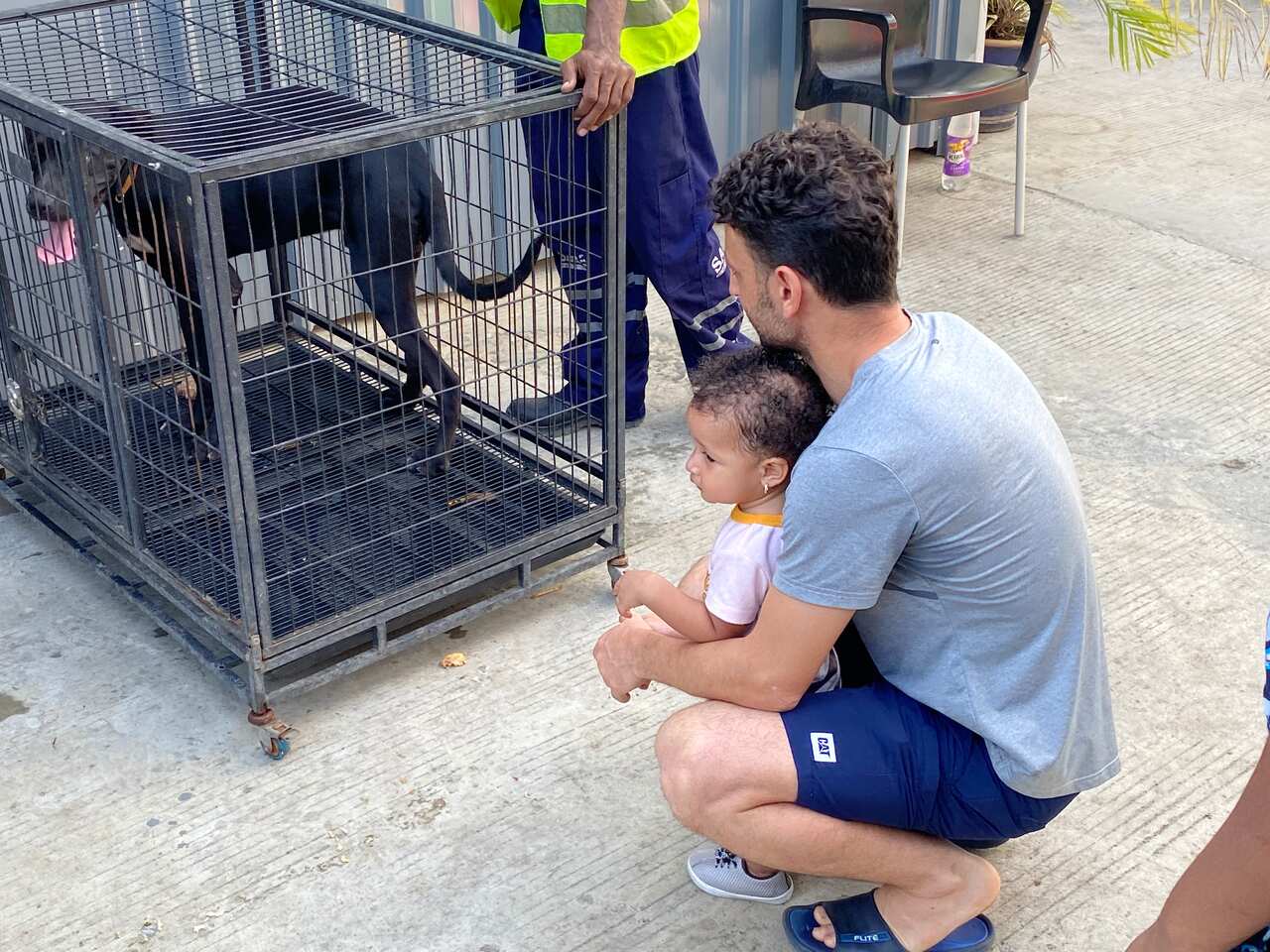 A man crouching down holding a baby and looking at a dog in a cage