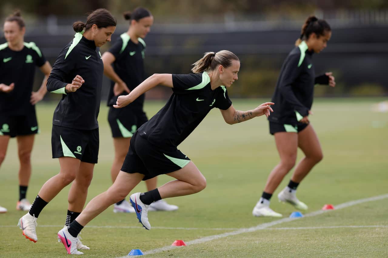Five players dressed in black and light green exercise clothes train on a grassed area with lines painted on it and coloured cones placed along the lines. Player Emily van Egmond is in the centre mid-run.