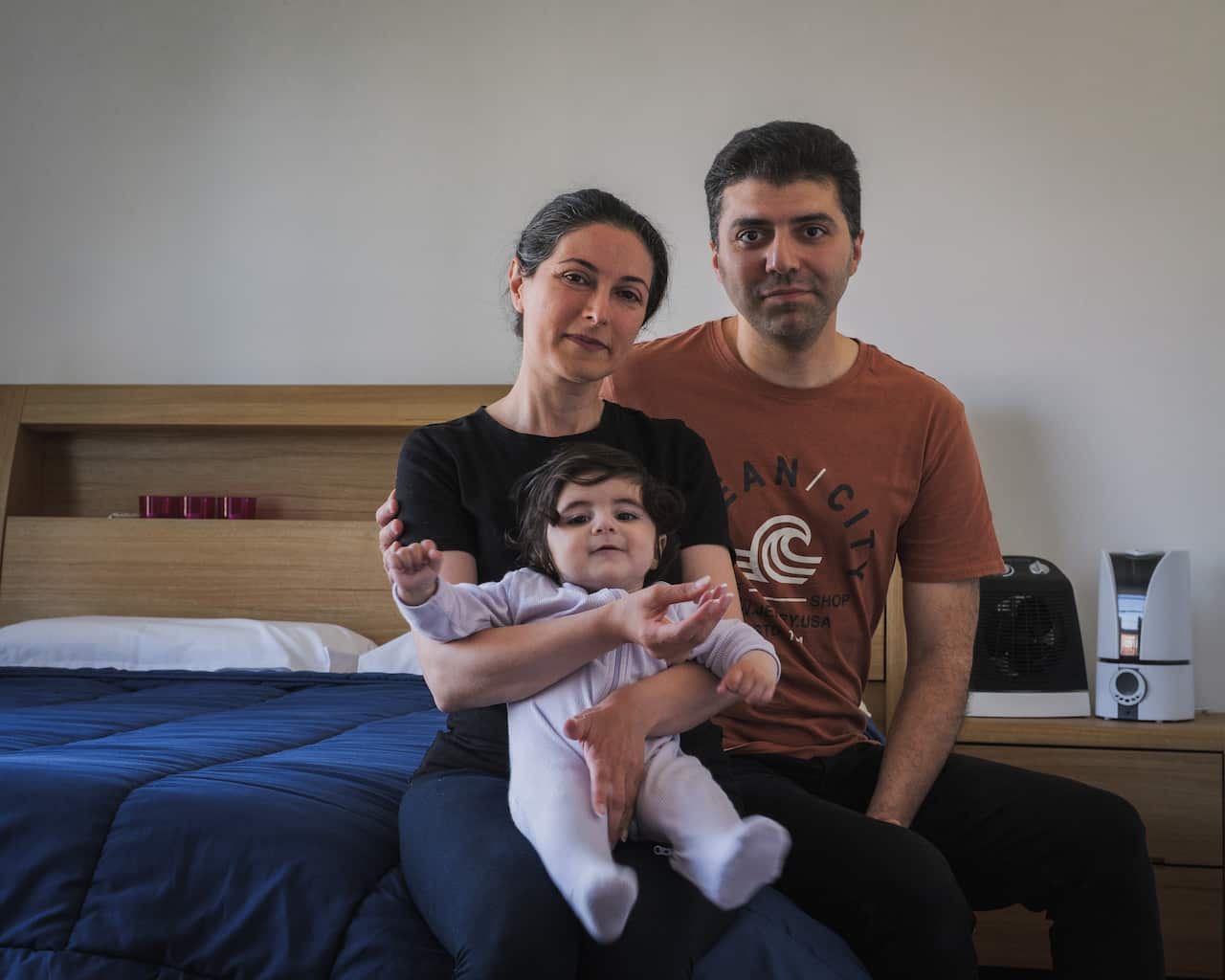 A family of three is seated on a bed, with the mother holding the child in her lap.