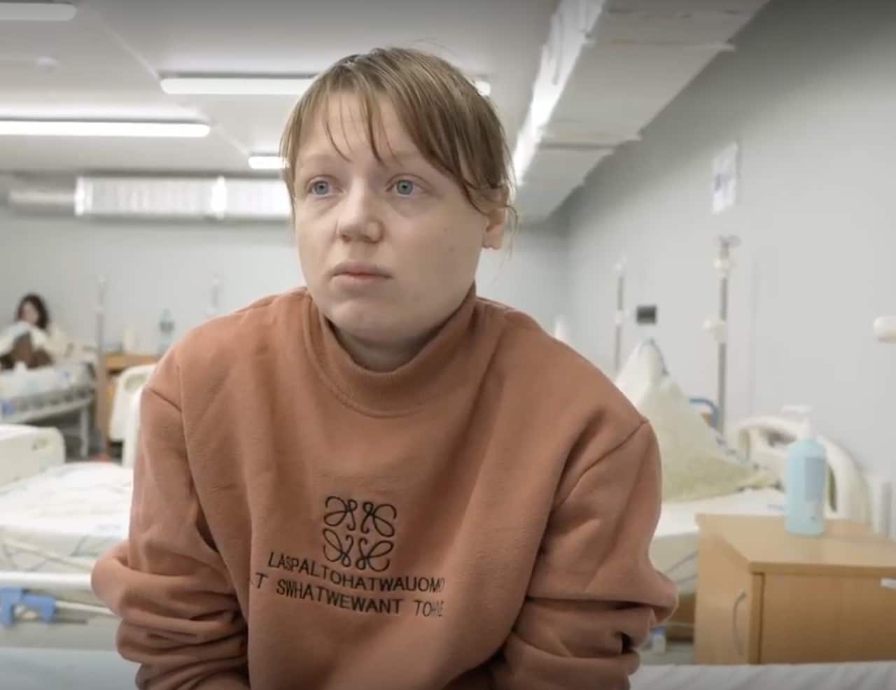 A woman with hair tied back wearing a brown top sits in a hospital ward with an anxious expression on her face.