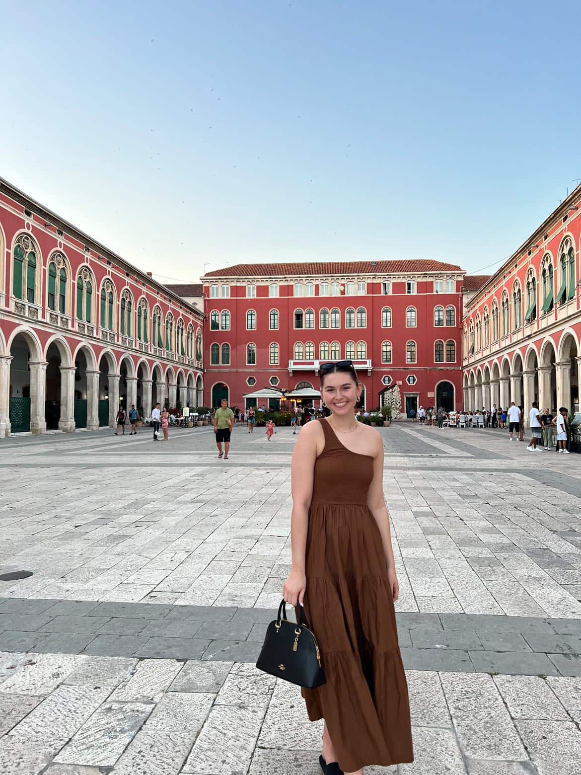 A smiling woman in a brown dress stands outside the Red Palace in Croatia. 