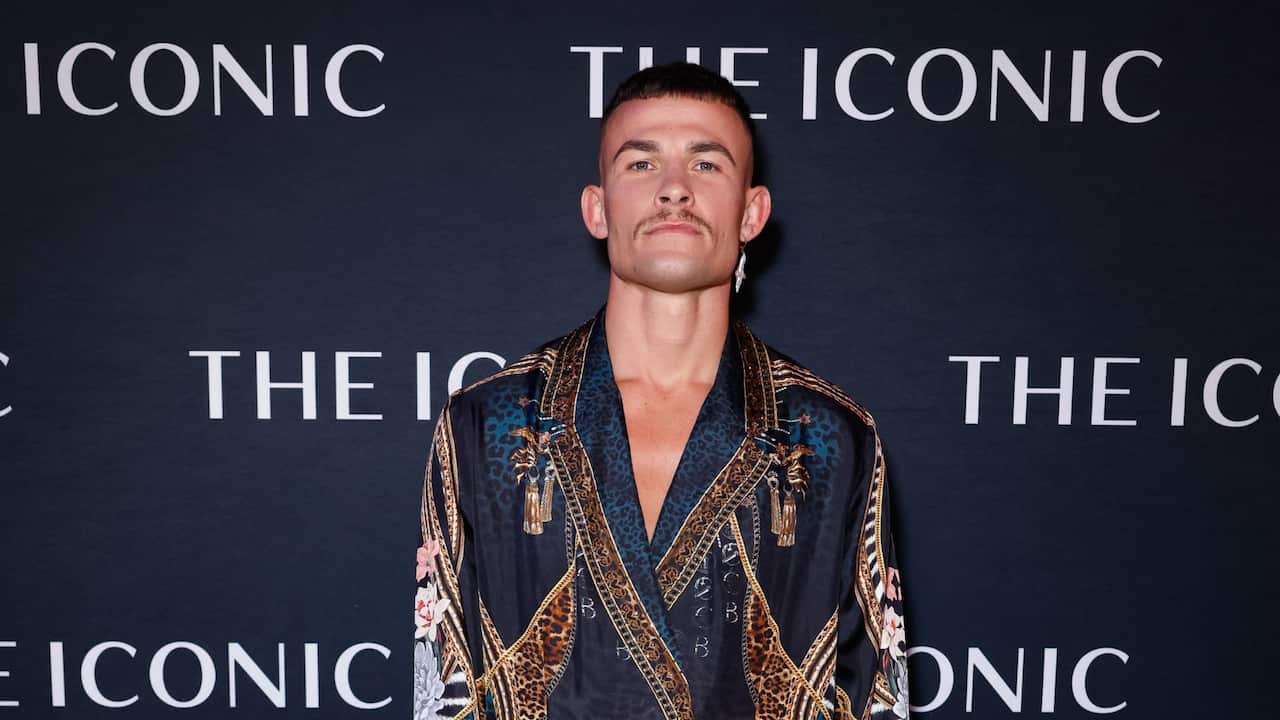 a young man in an expensive-looking patterned silk top stands in front of a background with THE ICONIC branding on it