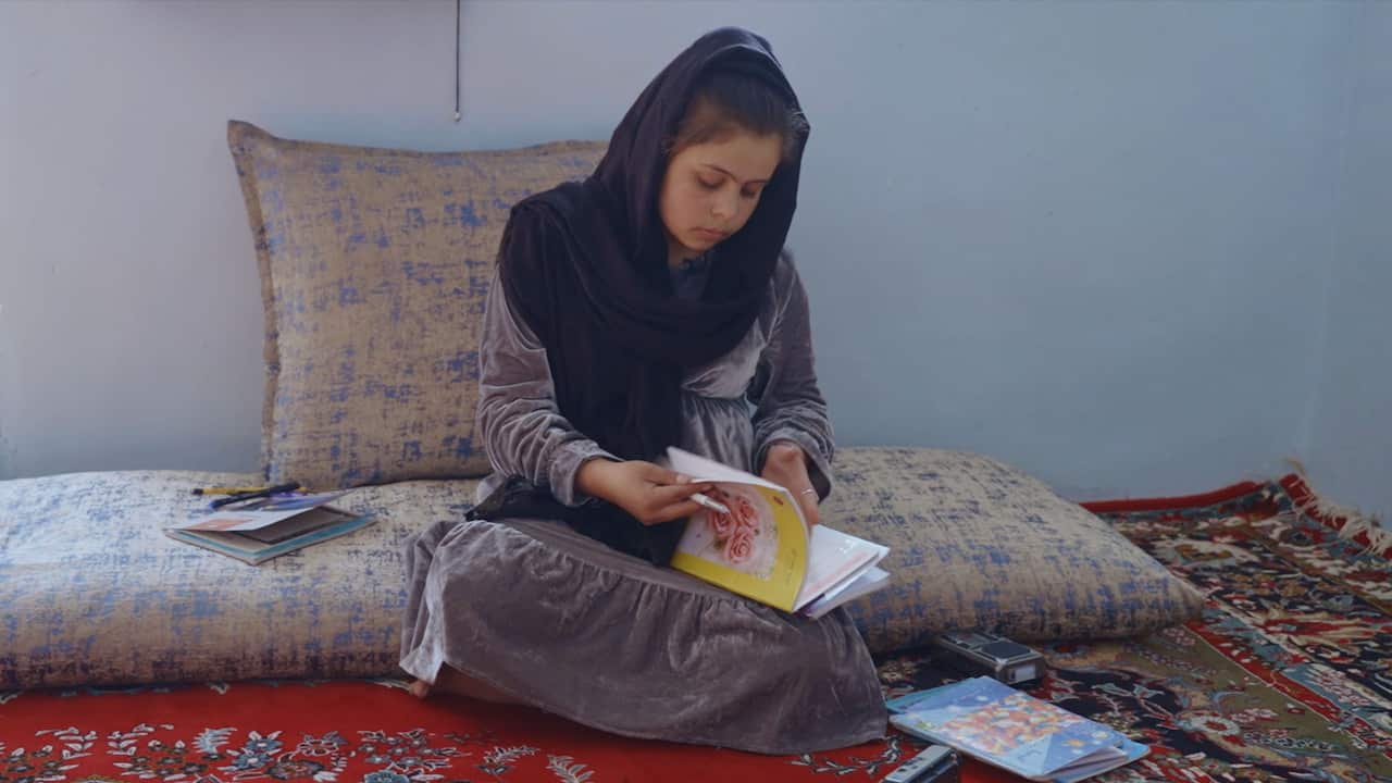 A teenage girl wearing a long grey dress and a black headscarf is seated on the floor covered with carpets and cushions with a book and a pen in her hands.