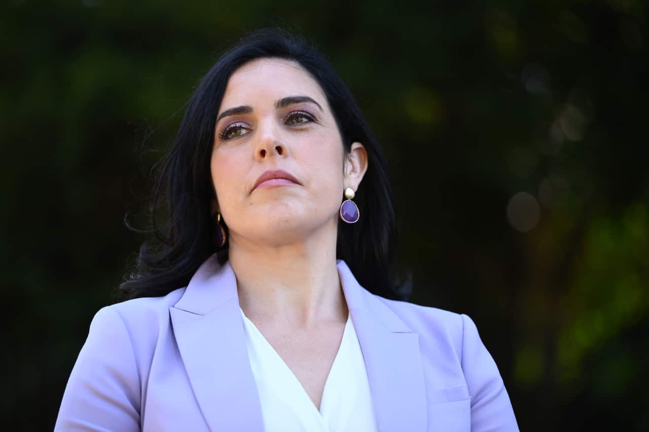 A woman in a purple blazer and white shirt looks forward with a serious expression on her face.