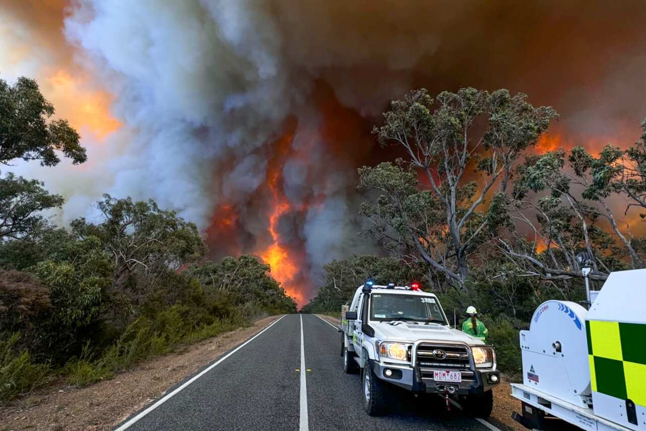 Smoke billows from an out-of-control bushfire in the background, with fire personnel vehicles on the road.