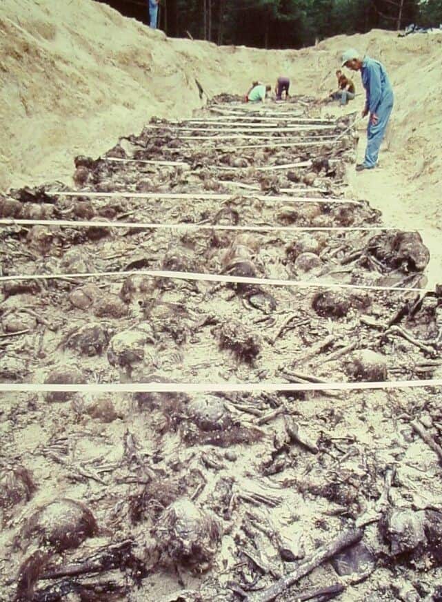 A mass grave filled with the decomposed and skeletal remains of dozens of human bodies, laid side by side in rows. Forensic investigators in protective clothing work at the far end of the grave, using tools to examine and document the site. The trench is surrounded by sand, and the scene is set in a wooded area.