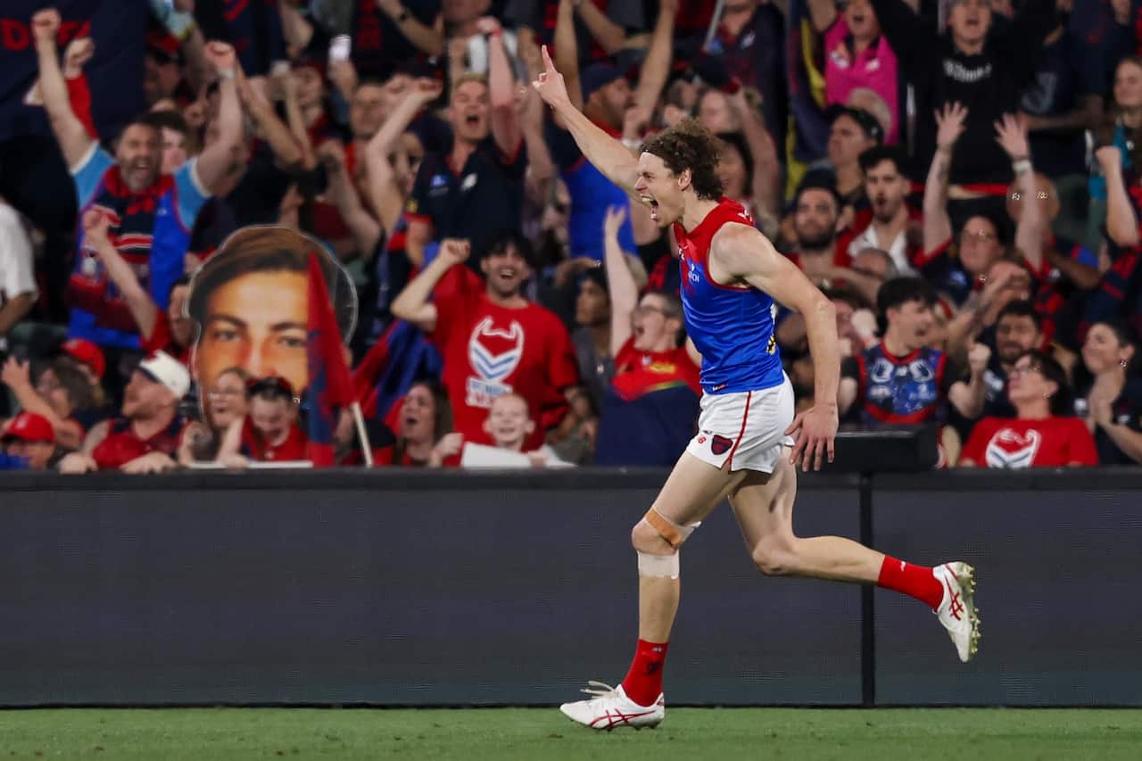 A man wearing a blue and red Aussie rules top runs and raises his arm in celebration on the field.