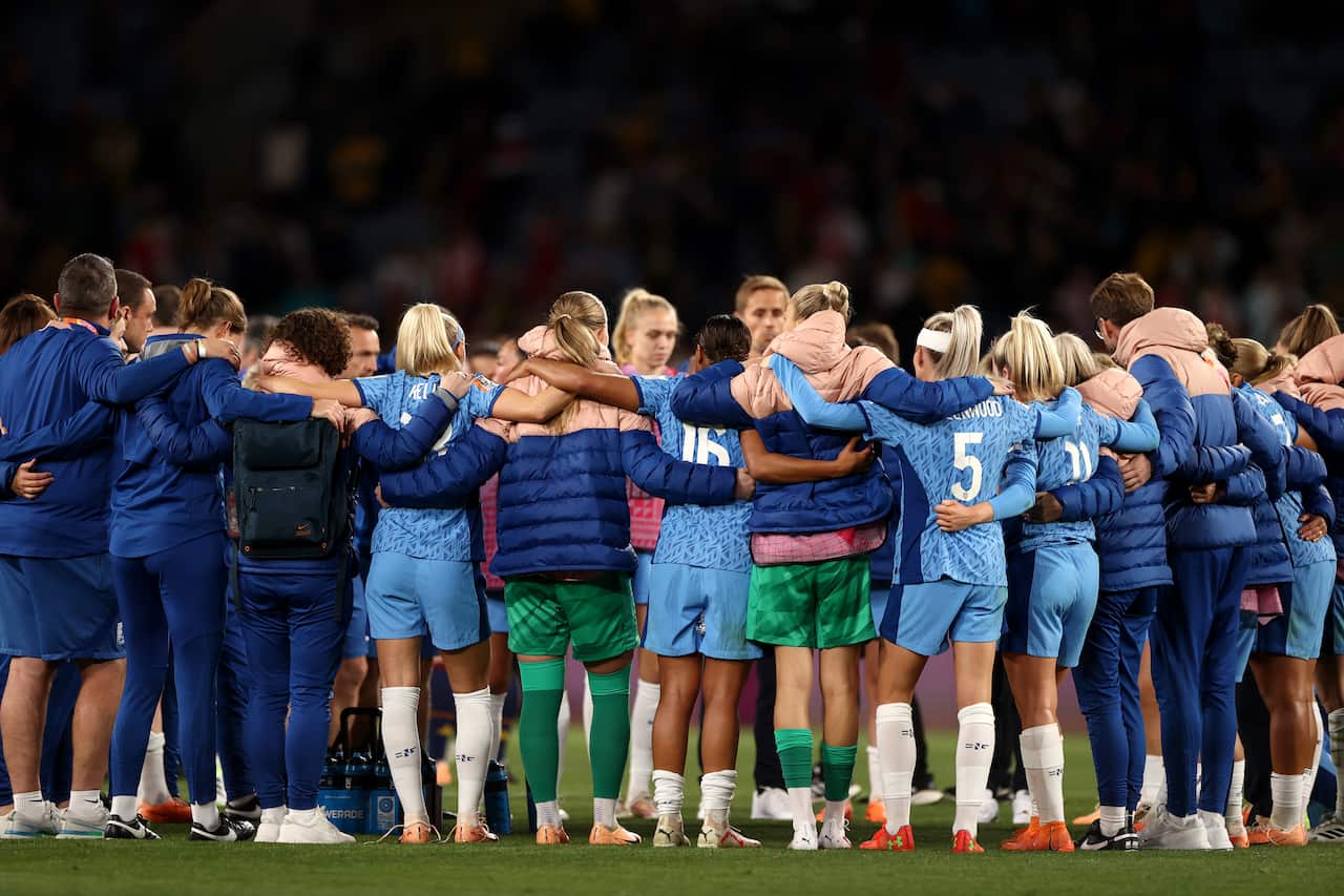 A group of football players in blue jerseys come together on the pitch, holding one another.