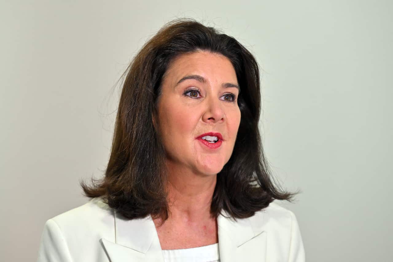 A woman with dark brown hair wearing a white blazer against a grey background.