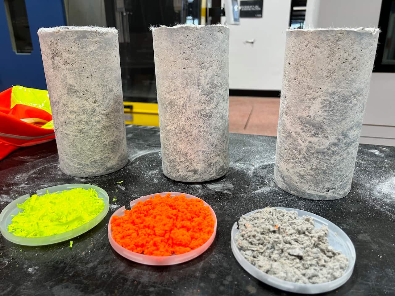 Three concrete cylinders stand behind dishes filled with coloured fibres.