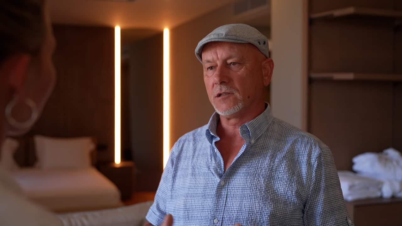 An older man wearing a light blue short-sleeved shirt and a grey flat cap speaks to another person in a hotel room.