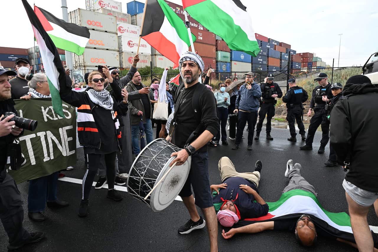 A man holding a drum stands next to two men lying on a road draped in Palestinian flags. People behind them wave Palestinian crowds and several police officers can be seen. 