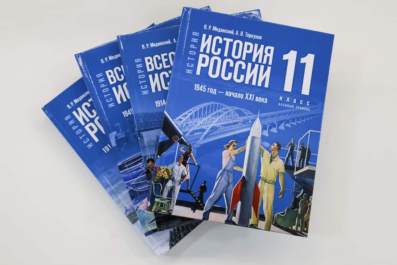Four Russian history textbooks stacked up together, with the one on the top showing an illustration of a man and a woman next to an aircraft bomb, other people standing around and the bridge that links Russia to annexed Crimea in the background. 