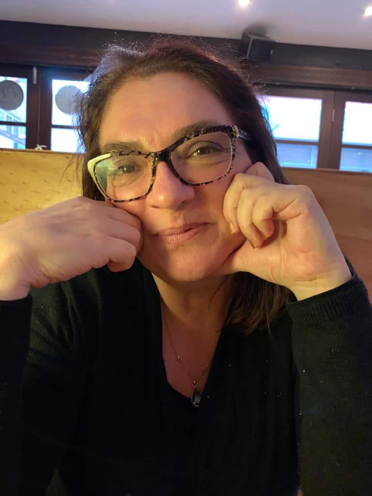 A woman in a black top and glasses smiles at the camera, her hands on her cheeks.
