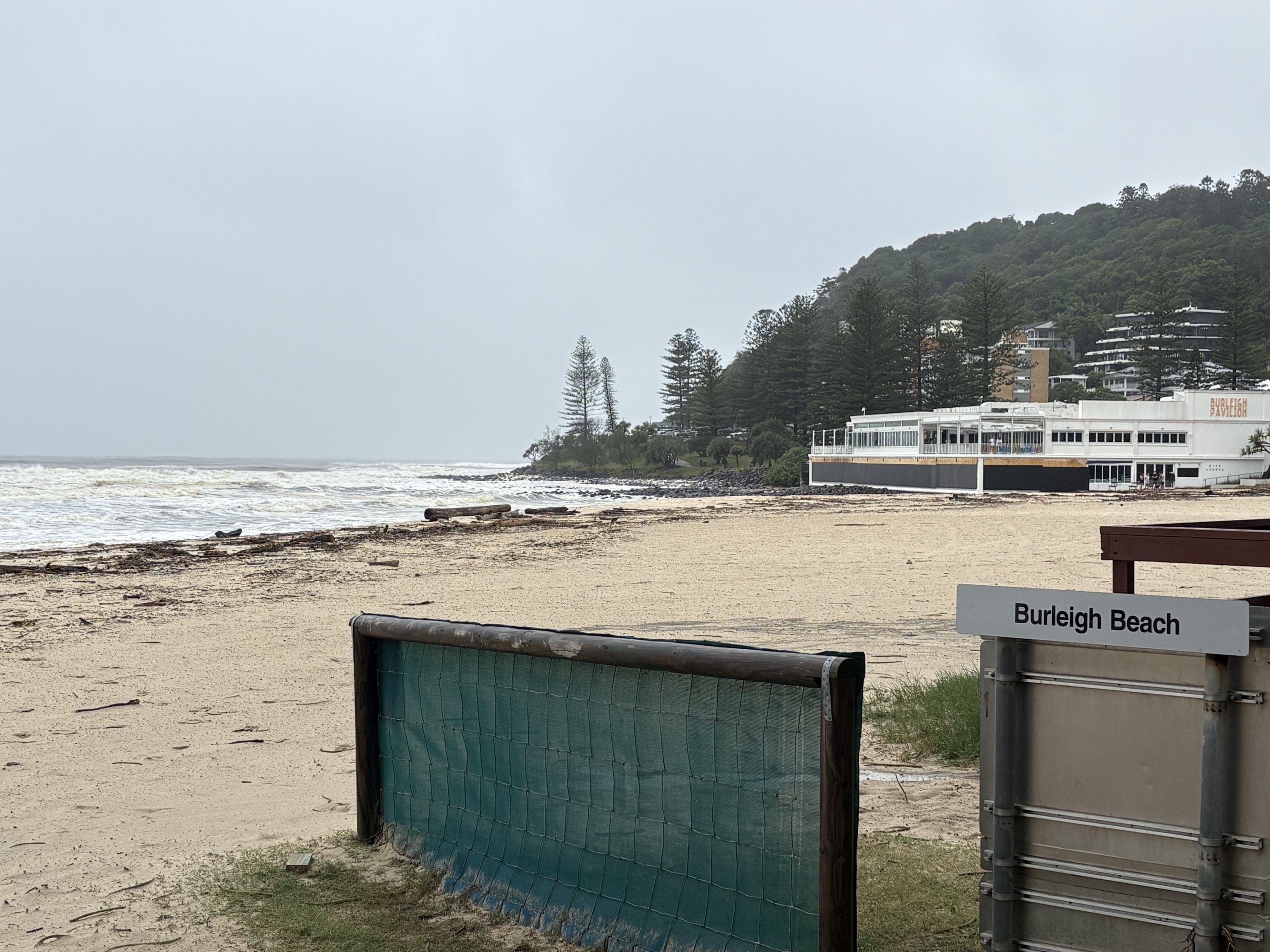 Tree branches and debris on the edge of the sand with a sign in the bottom right corner saying Burleigh Beach.