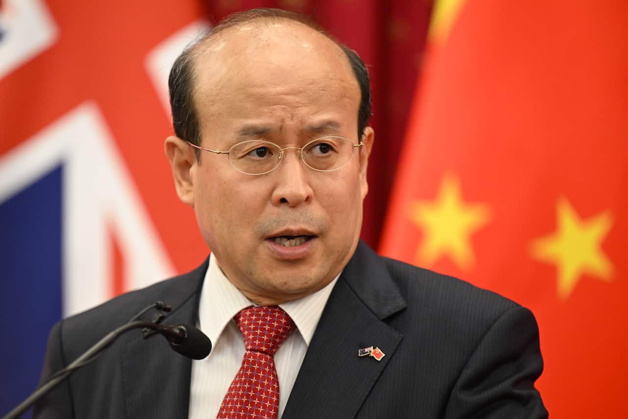 A man stands in front of a lectern, with Chinese and Australian flags blurred in the background.
