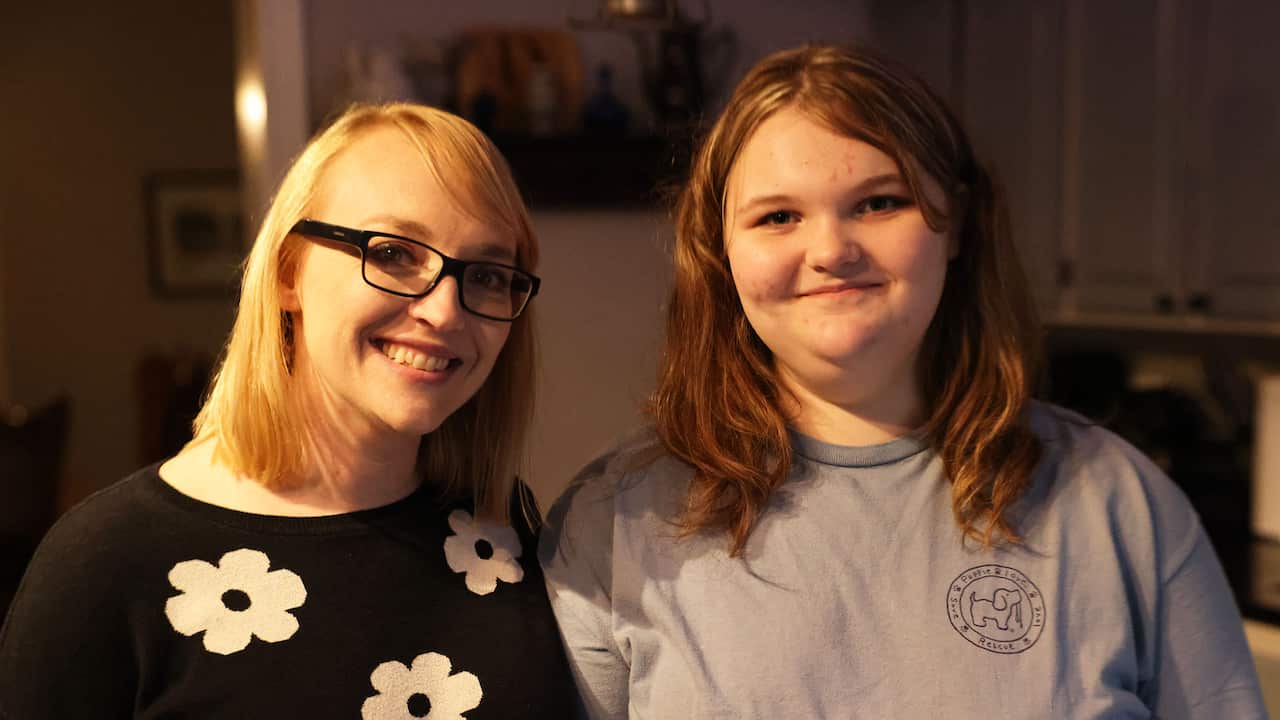A blonde woman in a black floral sweater and reading glasses stands next to a teenage girl in a grey t-shirt 