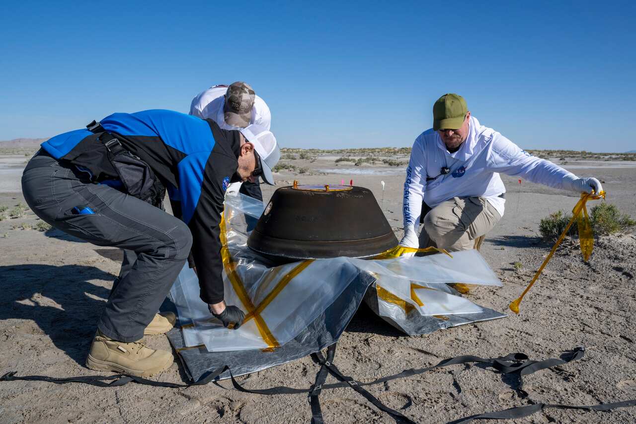 Three members of NASA circled around the OSIRIS-REx capsule that landed in the desert, preparing it for transport.