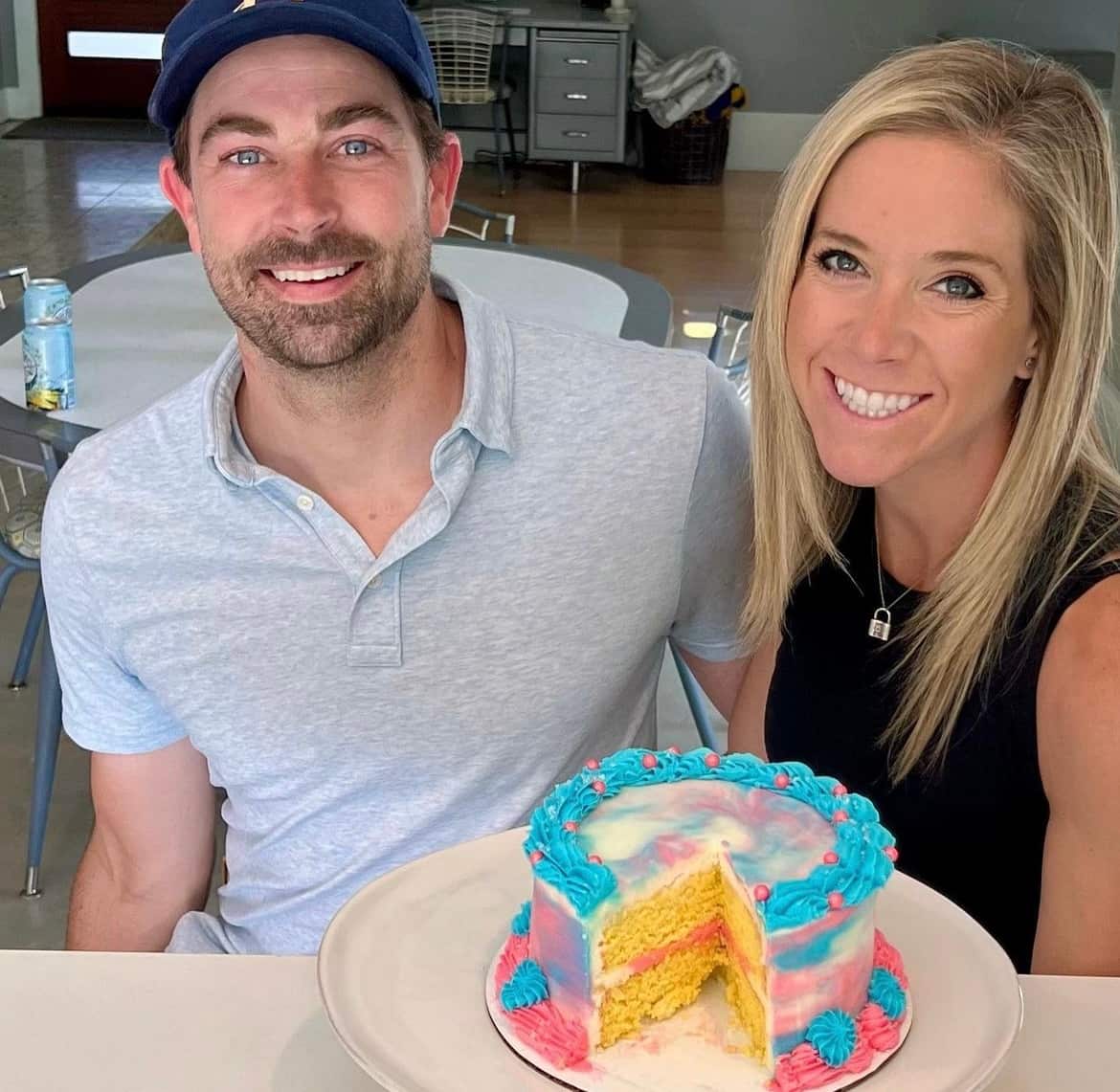 A man and a woman are smiling, with a cake with pink and blue icing placed before them on a table.