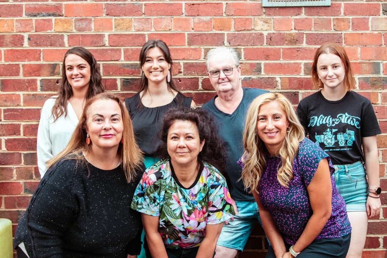 Seven women of various ages pose for a photo in front of a red brick wall.