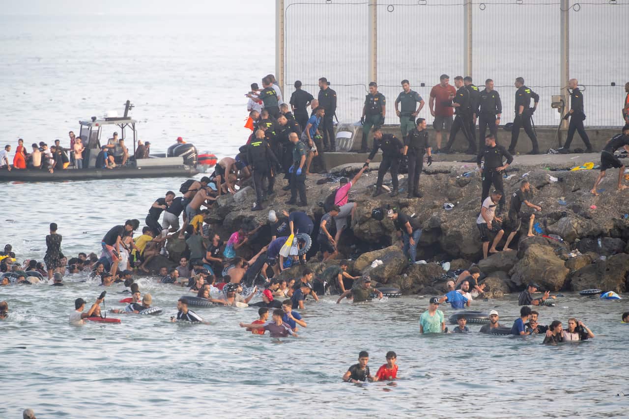 Dozens of migrants in the sea next to a rock breakwall. Uniformed police are standing on the breakwall, and a small boat carrying more migrants is in the background.