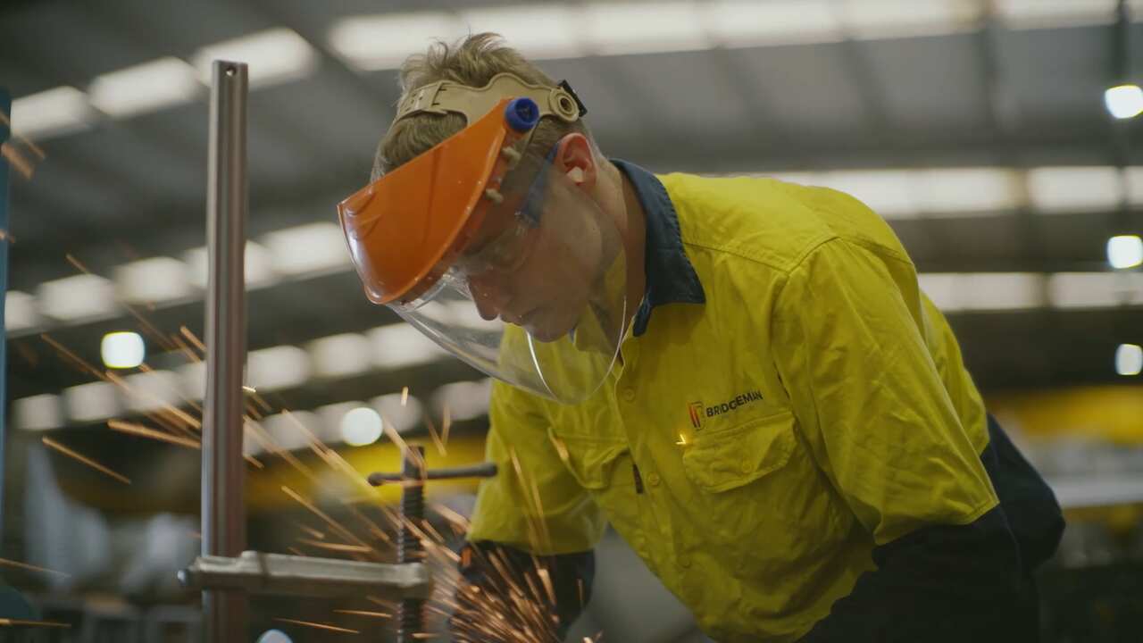 A man in yellow tradewear and protective helmet is grinding metal with a focussed face