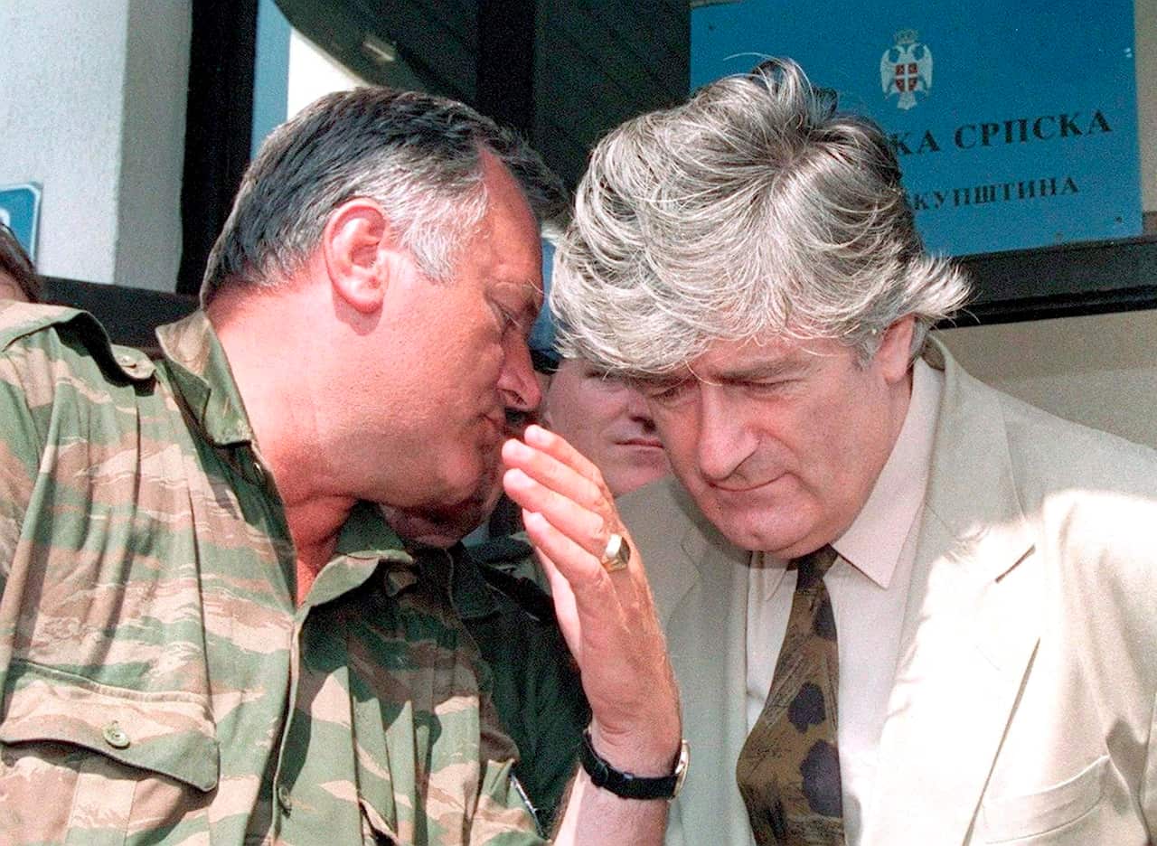 Two middle aged men having a conversation. The man on the left is wearing military camouflage, the man on the right a pale green suit jacket, white shirt and brown splotched tie. 