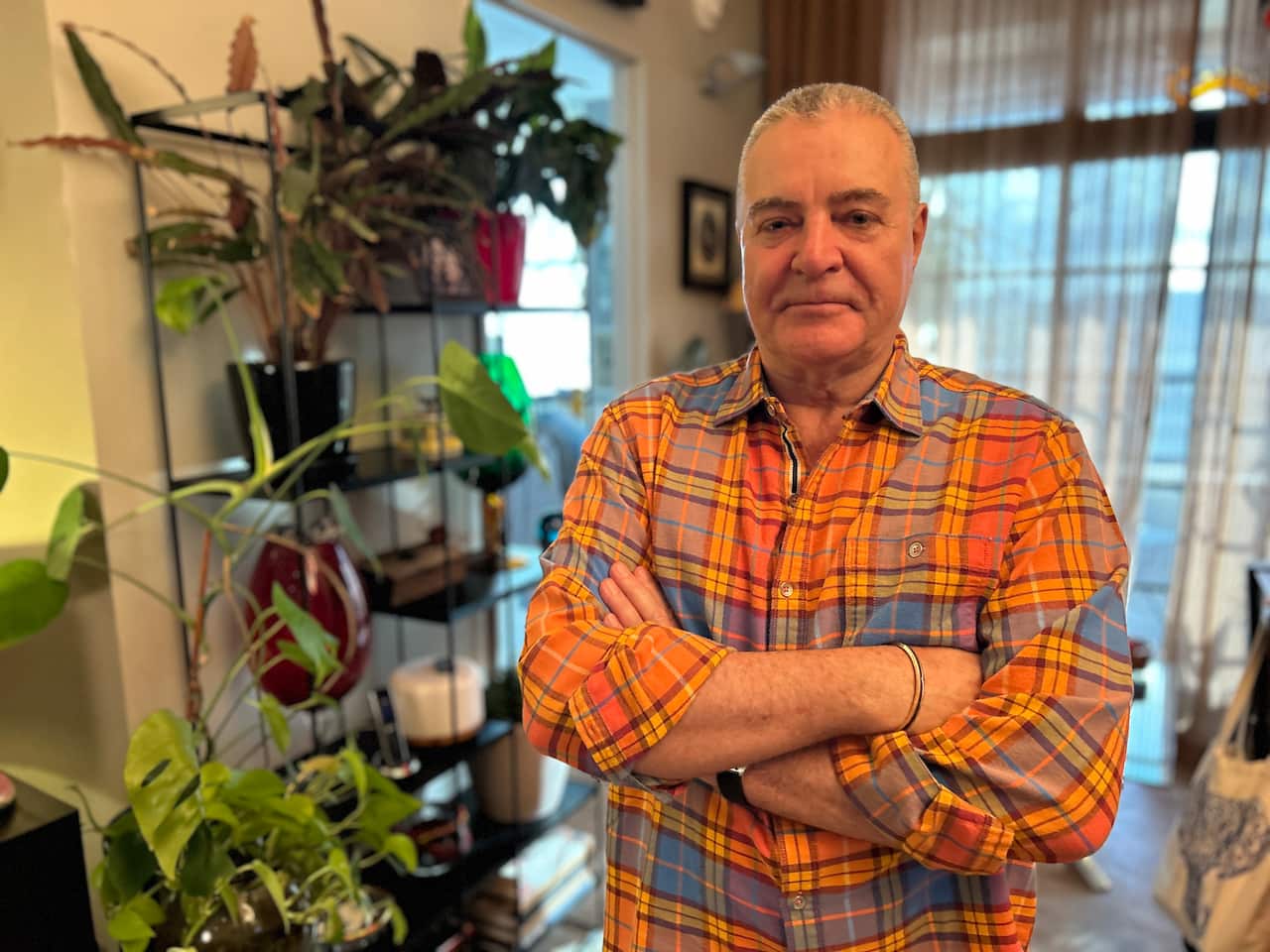 A man with grey cropped hair wearing a brightly coloured checked shirt stands in his home with his arms folded next to a plant.