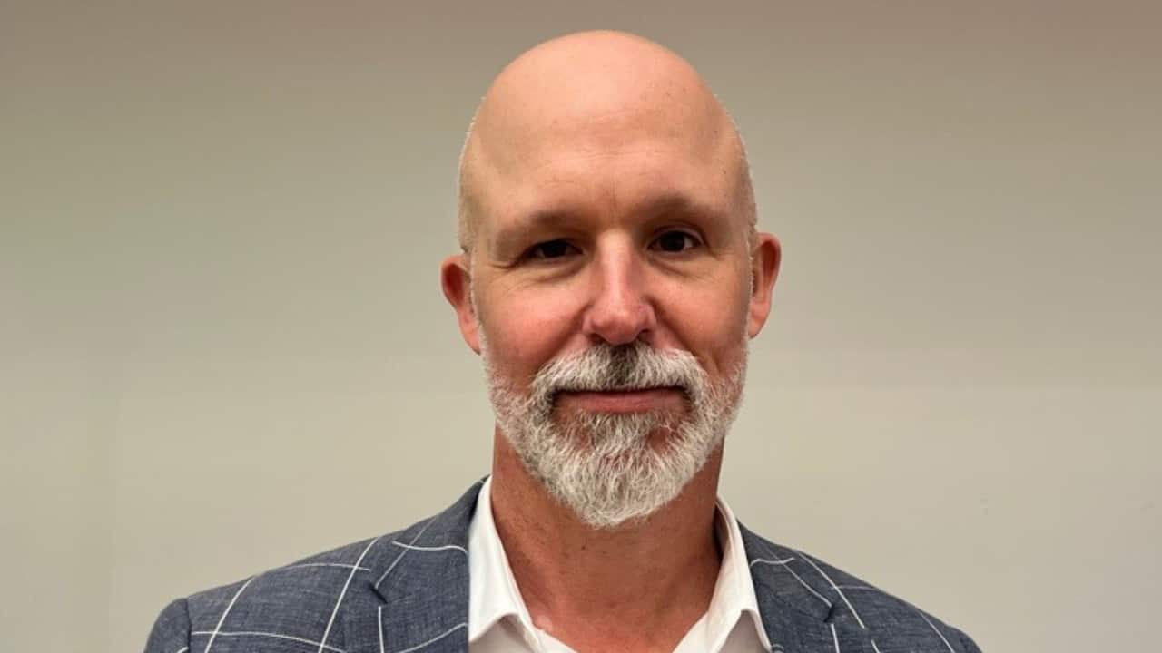 A bald man with a white goatee beard. He is wearing a grey checked suit jacket