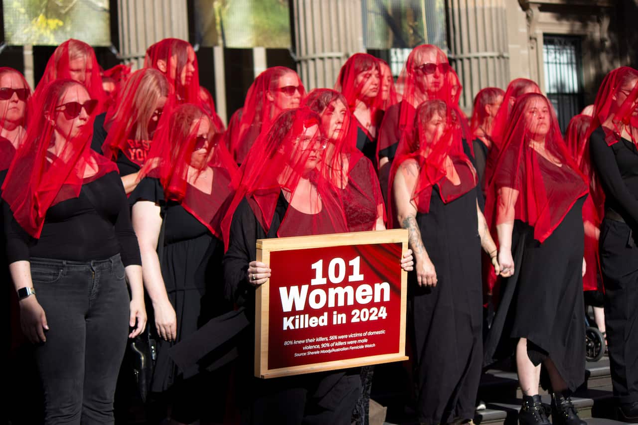 Around a dozen women dressed in black with red veils over their faces stand on steps in front of a building. One holds a sign reading, 101 Women Killed in 2024