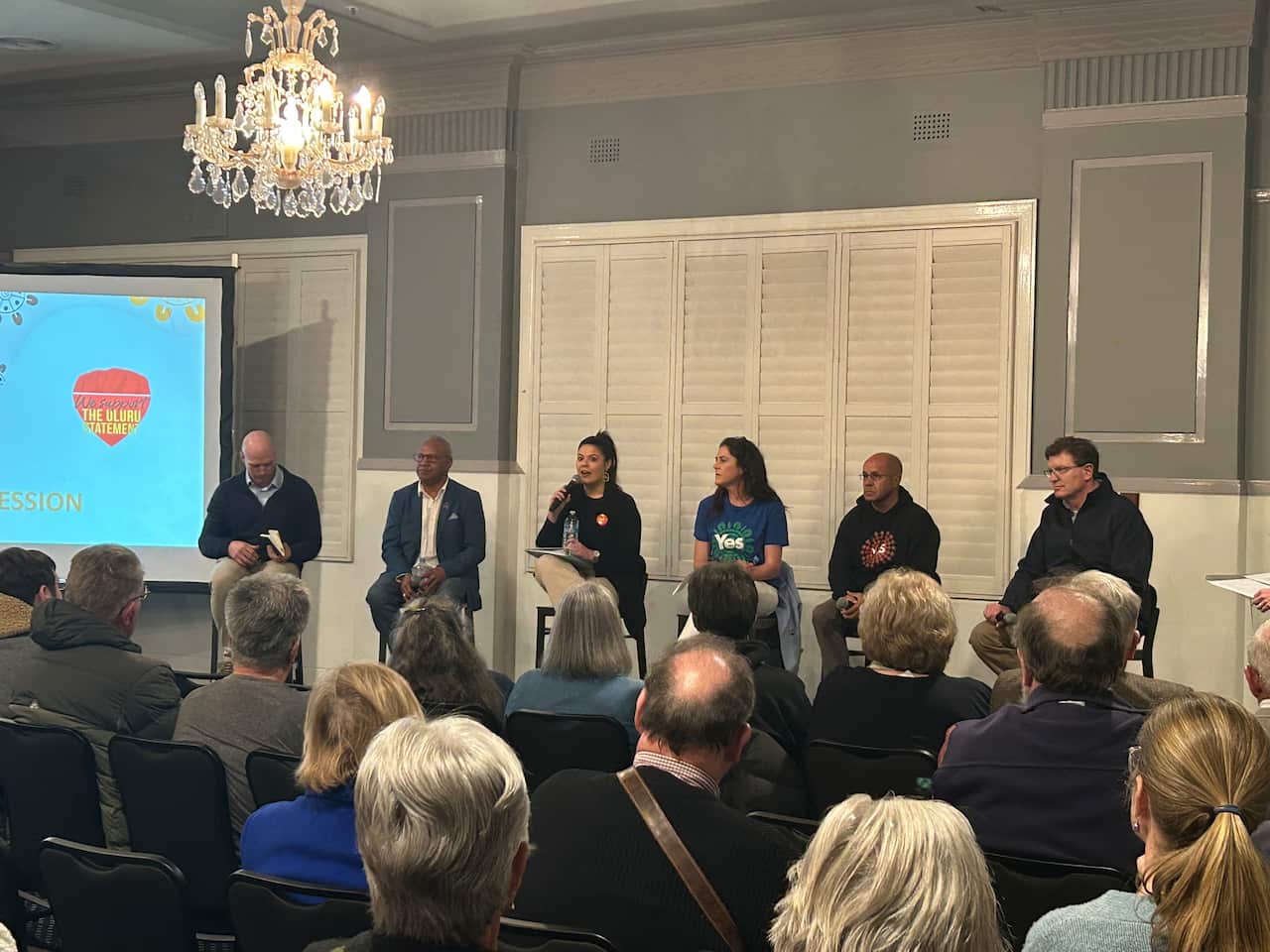 Six people sitting on high chairs at the front of a room full of people sitting in chairs