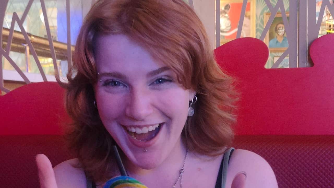 a close up of a young girl with red hair and an eyebrow piercing sits in a red booth smiling