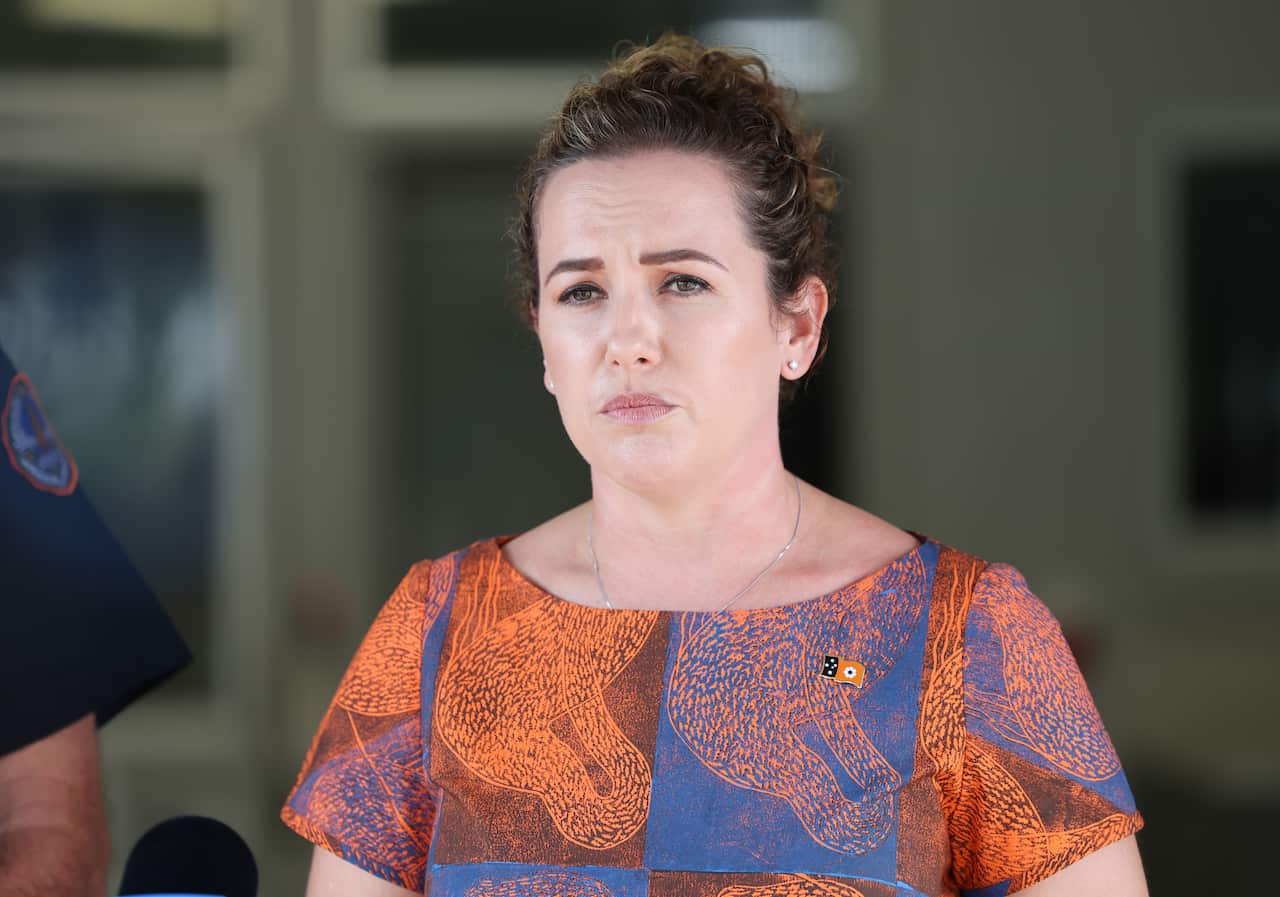 A woman with brown curly hair wearing an orange and blue dress looks forward with a serious expression on her face. 