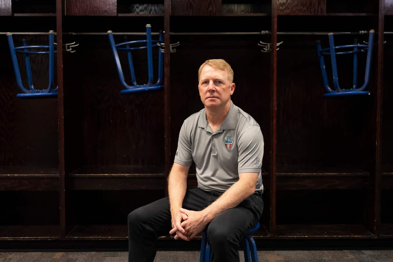 A man wearing black trousers and a grey polo shirt sitting in front of lockers