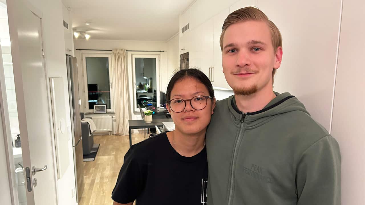 A young woman wearing prescription glasses and a young man pose for a photo, with the interior of their apartment visible in the background