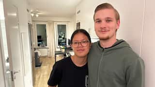 A young woman wearing prescription glasses and a young man pose for a photo, with the interior of their apartment visible in the background