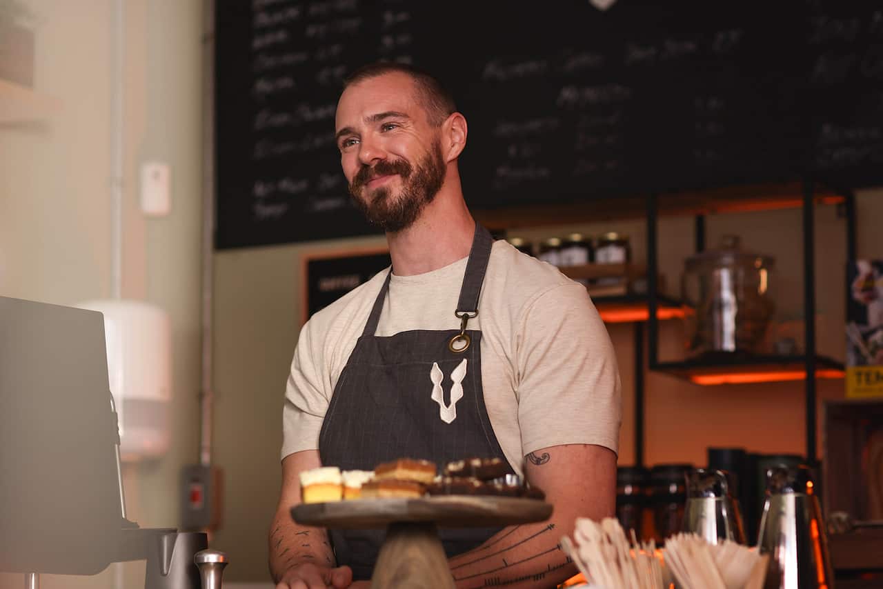 A smiling man in an apron stands in a cafe. 