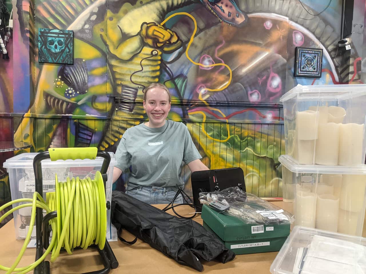 A young woman sits behind a table filled with items including an extension cord on a wind up spool, camping items and plastic tubs full of large decorative candles.