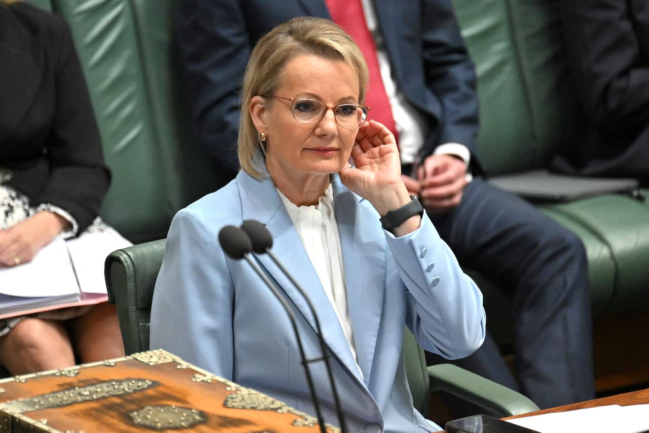 Sussan Ley wearing a blue coat, standing in a room with other people sitting on green seats.