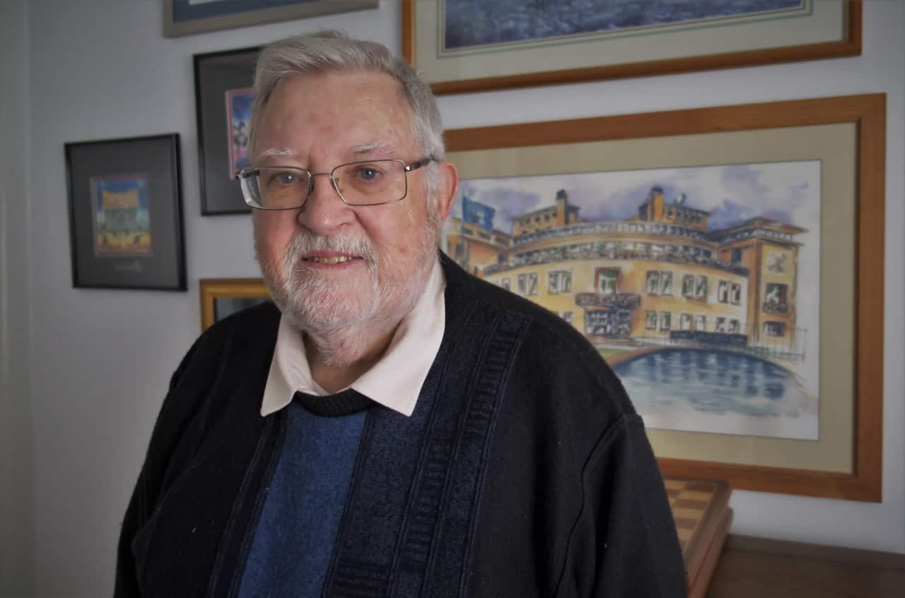An older man with white hair, a trimmed beard, and glasses stands indoors, smiling softly. He is wearing a dark sweater over a blue shirt with a white collar. Behind him is a wall filled with framed artwork, including a large watercolor-style painting of the International Criminal Tribunal for the former Yugoslavia building, featuring yellow tones and curved shapes beside a reflecting pool.