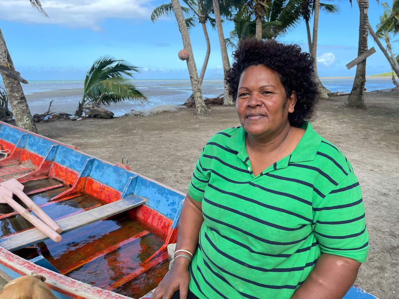 A woman sits on the side of a small wooden boat with water in it, which sits on wet sand in front of palm trees and the ocean in the background.