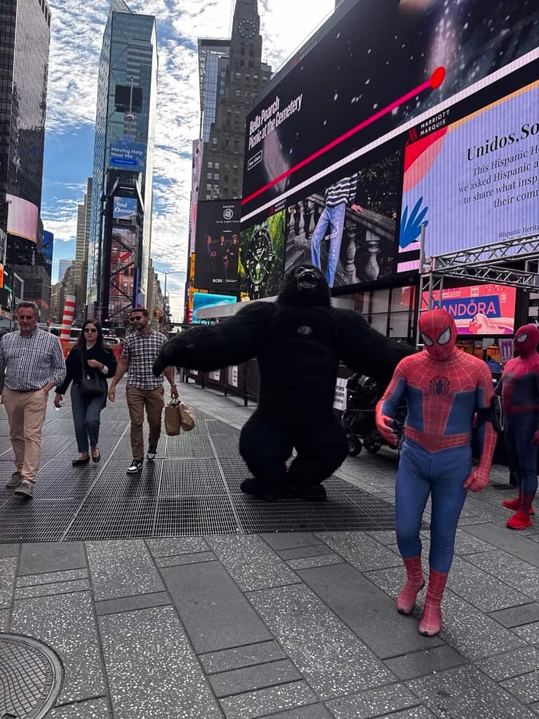 Times Square pedestrian area with two people in Spider-Man costumes and a person in a gorilla suit, as pedestrians walk past large digital billboards and skyscrapers.