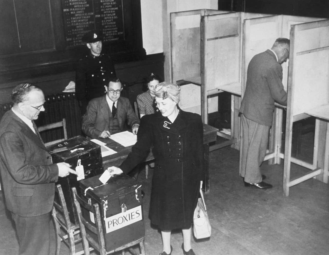 A black and white photo showing a woman in a black dress depositing a ballot paper into a box labelled "proxies". 