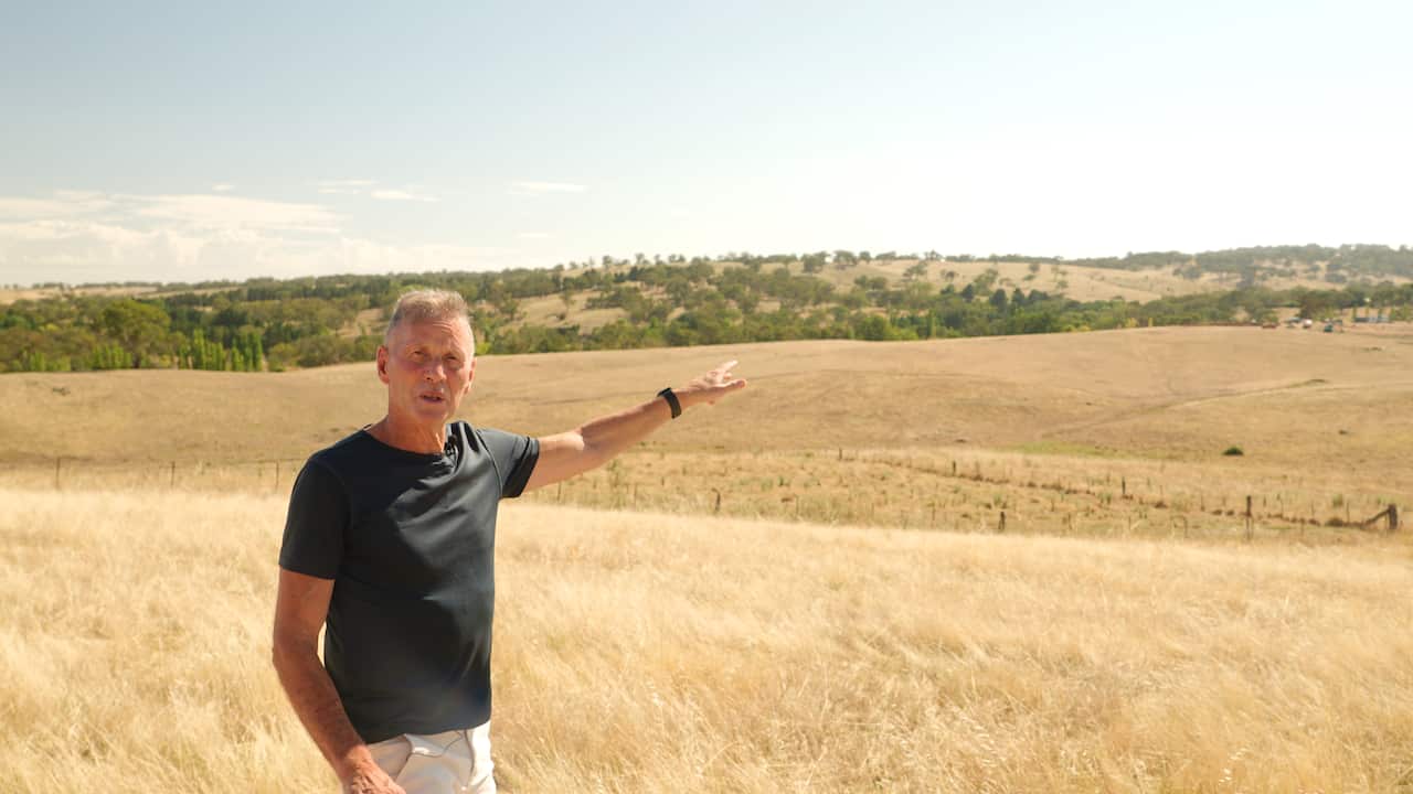 A middle-aged man with short grey hair and a black T-shirt points down toward a rural property where construction is underway, surrounded by dry grass and open landscape.