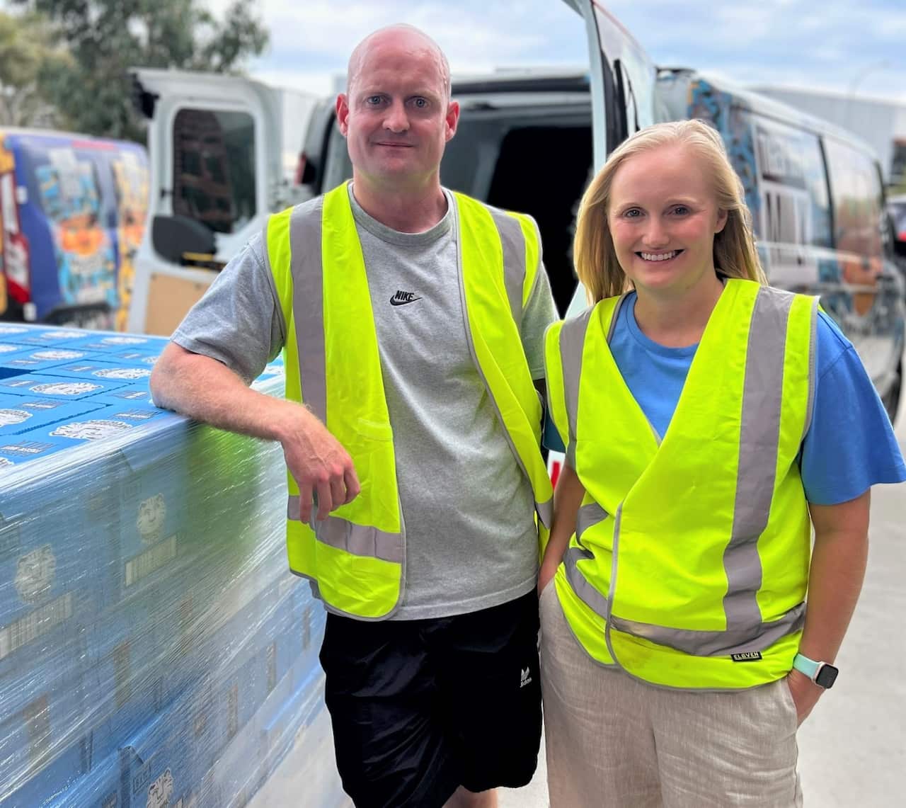 A man and a woman in hi-vis yellow vests standing together. The man is leaning on a pallet. A van with open back doors is in the background