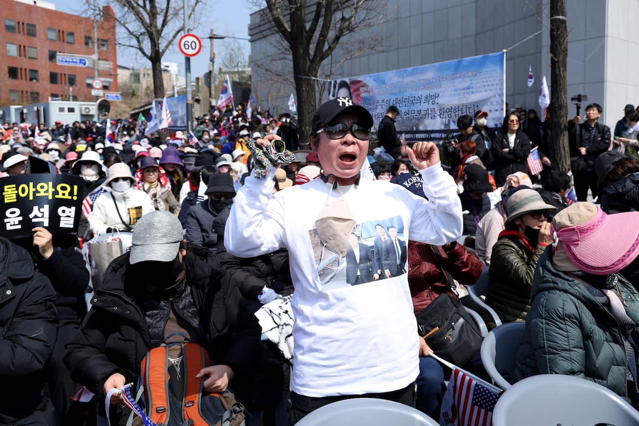 A woman wearing a black New York Yankees cap and a white shirt stands with her arms aloft in a crowd of protestors.
